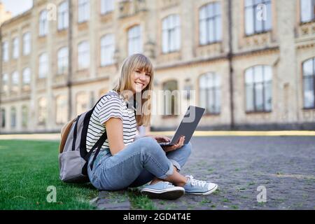 Jeune femme blonde caucasienne mignonne étudiante sur le campus de l'université, assise sur l'herbe verte avec sac à dos et ordinateur portable de navigation sur Internet bonne émotion regardant directement à l'appareil photo. Photo de haute qualité Banque D'Images