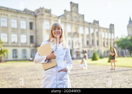 Portrait d'un jeune étudiant en médecine blonde avec un ordinateur portable debout à l'extérieur portant une robe médicale blanche. Les stagiaires ou le médecin étudiant du campus universitaire regardent la caméra. Concept éducatif Banque D'Images