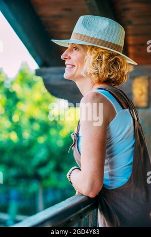 Portrait d'une jeune femme blonde de race blanche mature dans ses années 50 portant un chapeau sur le porche en bois d'une maison de campagne. Concept de style de vie. Banque D'Images