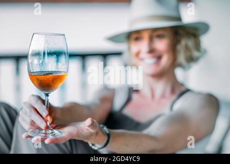 Jeune femme blonde de race blanche mature dans ses années 50 portant un chapeau et appréciant un verre de vin sur le porche d'une maison de campagne. Concept de style de vie. Banque D'Images