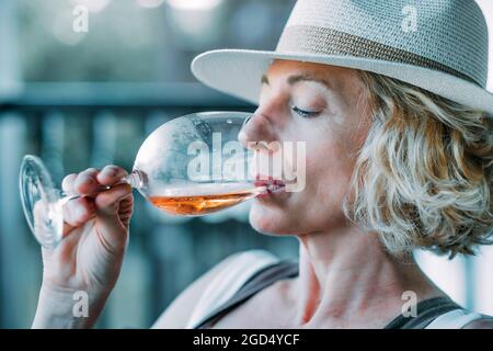 Jeune femme blonde de race blanche mature dans ses années 50 portant un chapeau et goûtant un verre de vin sur le porche d'une maison de campagne. Concept de style de vie. Banque D'Images