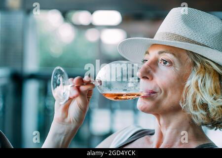 Jeune femme blonde de race blanche mature dans ses années 50 portant un chapeau et goûtant un verre de vin sur le porche d'une maison de campagne. Concept de style de vie. Banque D'Images