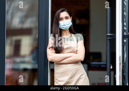 Ouverture du café après la quarantaine Covid-19. Jeune femme arabe en tablier et masque de protection avec bras croisés regardant la caméra attendant les clients près de fron Banque D'Images