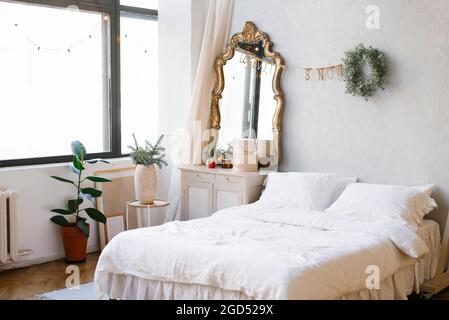 Lit double avec draps et oreillers blancs dans la chambre décorée pour Noël et le nouvel an. Couronne de Noël près du miroir de la commode Banque D'Images