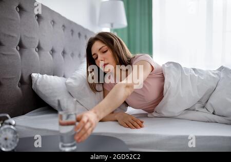 Femme de race blanche millénaire prenant un verre d'eau minérale claire de la table de chevet après s'être réveillée au lit à la maison. Une jeune femme qui restait hydratée, qui prenait Banque D'Images
