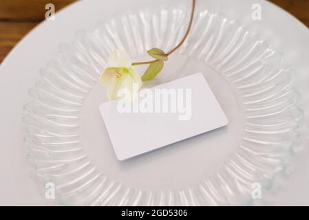 Une maquette blanche vierge d'une carte d'invité sur une plaque de verre et d'une fleur blanche délicate . Pour un banquet de fête, un mariage ou une fête d'anniversaire Banque D'Images