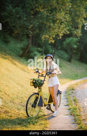 Une jeune belle femme tient un vélo avec des fleurs de verveine jaune dans un panier lors d'une randonnée en plein air Banque D'Images