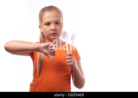 Jolie petite fille caucasienne tenant des couverts en plastique sur un fond blanc et montrant son pouce vers le bas. Vaisselle jetable, concept de recyclage en plastique. Banque D'Images