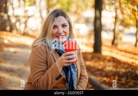 Belle femme blonde d'âge moyen souriante portant un manteau et une écharpe beige, assise dans le parc de la ville le jour d'automne ensoleillé, appréciant sa tasse de café à emporter. L Banque D'Images