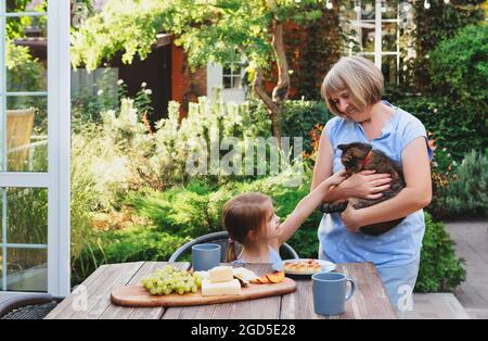 Petite fille mignonne assise à table et prenant le petit déjeuner dans le jardin de la maison de campagne, chat de petting assis dans les bras de grand-mères, petite-fille de dépenser Banque D'Images