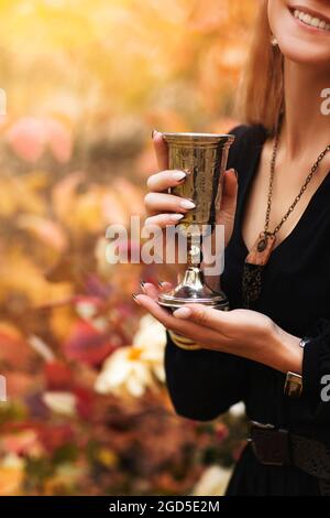 Coupe courte de jeune femme gaie en robe noire tenant le verre métallique vintage dans les mains tout en se tenant dans le parc d'automne avec des feuilles d'arbre colorées sur la ba Banque D'Images