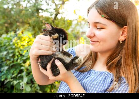 Photo courte d'une jeune fille en robe rayée bleue tenant un adorable lapin moelleux avec les mains accrochant doucement à sa poitrine debout à la campagne estivale Banque D'Images