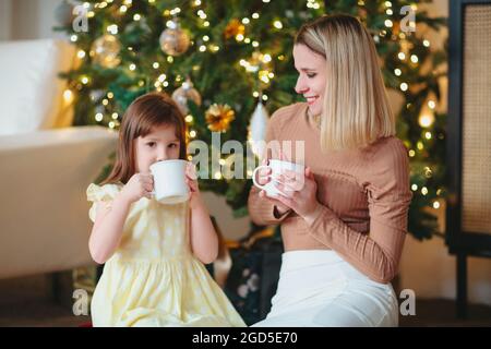 Famille heureuse, mère et petite fille buvant caçao chaud, assis près de l'arbre ?hristmas décoré, maman et enfant passant du temps ensemble à la maison dans la durée Banque D'Images