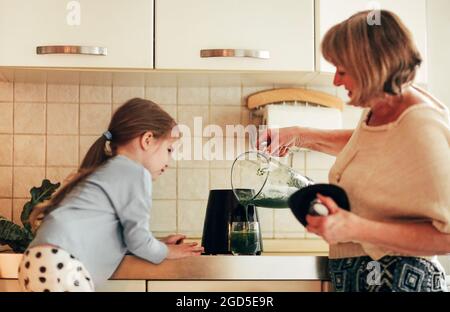 Grand-mère aimant cuisiner avec la petite-fille à la maison, en utilisant le mélangeur pour mélanger les légumes, petite fille apprenant à utiliser des appareils avec grand Banque D'Images