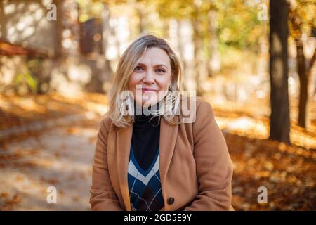 Heureuse femme d'âge moyen qui profite de l'automne à l'extérieur, vêtue de vêtements décontractés et d'un manteau beige debout dans un parc jaune, regardant de côté et souriant gaieté Banque D'Images