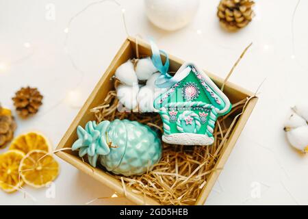 Boîte cadeau de Noël avec maison en pain d'épice et bougie. Souvenir de fête en argile polymère. Surprise du nouvel an, cônes et oranges séchées sur table blanche. Composition pour la conception de cartes postales de vœux. Banque D'Images