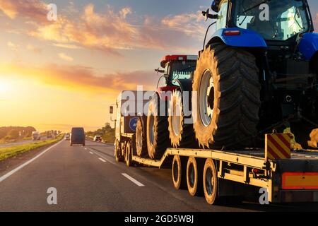 POV lourd camion industriel semi-remorque plate-forme de transport de deux gros tracteur agricole moderne sur la route commune au coucher du soleil ciel lever du soleil Banque D'Images