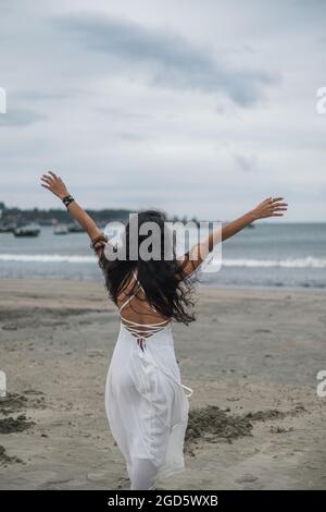 Belle femme en robe blanche marchant sur la plage. Mains sur le côté. Vue arrière. Fond marin. Cheveux longs et bouclés noirs. Vent dans ses cheveux. Banque D'Images