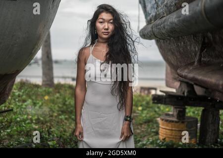 Belle jeune femme mongol en robe blanche debout entre deux vieux bateaux en bois. Brunette cheveux longs bouclés. Pensif. Banque D'Images