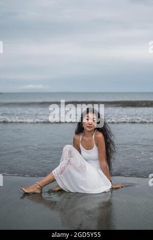 Charmante jeune femme mongol en robe blanche assise sur la plage. Cheveux longs et bouclés noirs. Regarder l'appareil photo avec l'espace de copie. Vue sur l'océan. Banque D'Images