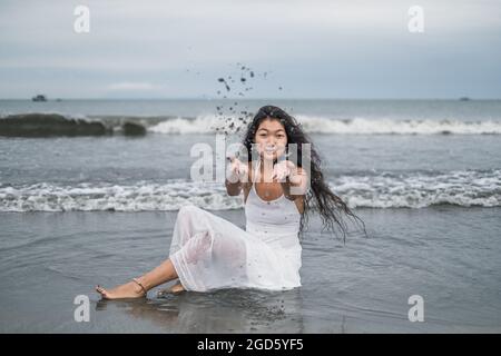 Charmante jeune femme mongol en robe blanche assise sur la plage et jouant avec le sable. Lancer l'envoi. Cheveux longs et bouclés noirs. Romantique. Vue sur l'océan Banque D'Images