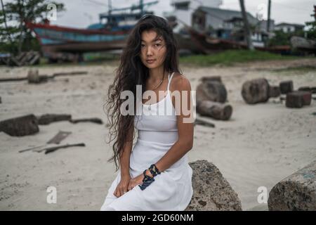 Charmante jeune femme asiatique en robe blanche assise sur la pierre et regardant l'appareil photo avec espace de copie. Brunette cheveux longs bouclés. Portrait romantique. Banque D'Images