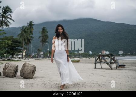 Belle jeune femme asiatique en robe blanche marchant sur la plage. Fond de montagne. Cheveux longs et bouclés noirs. Vent dans ses cheveux. Photo romantique. Banque D'Images