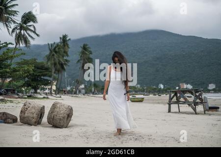 Charmante jeune femme en robe blanche marchant sur la plage et regardant vers le bas. Fond de montagne. Cheveux longs et bouclés noirs. Vent dans ses cheveux. Banque D'Images