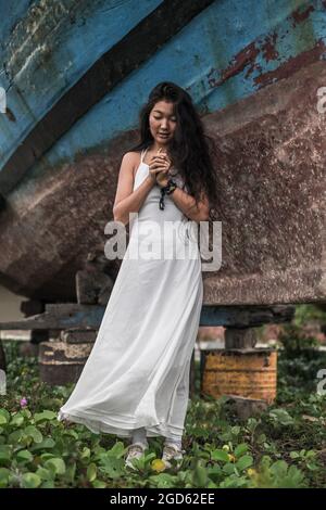 Charmante jeune femme mongol en robe blanche regardant ses mains serrées à la poitrine. Cheveux longs Brunette. Photo de haute qualité Banque D'Images