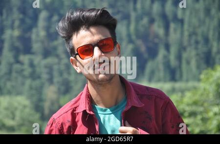 Gros plan d'un jeune homme du nord de l'Inde posant à l'extérieur avec porter une chemise rouge et des lunettes de soleil rouges avec regarder l'appareil photo Banque D'Images