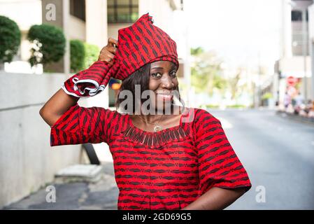 femme africaine élégante et tendance portant un tissu rouge lanières une écharpe sur sa tête tout en souriant dans la rue à la lumière du jour Banque D'Images