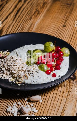 Bol à smoothie avec groseilles à maquereau et raisins de Corinthe. Baies fraîches, flocons d'avoine, pistaches sur plaque noire sur table rustique en bois, gros plan Banque D'Images