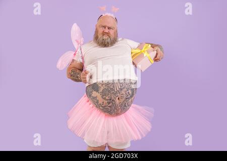 Drôle d'homme de plumpe barbu en costume de fée avec boîte cadeau grimaces sur fond violet Banque D'Images