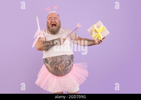 Un homme obèse excité en costume de fée tient une boîte cadeau sur fond violet Banque D'Images