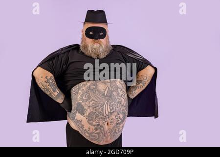 Homme confiant avec un excès de poids en costume Zorro tient les mains à la taille sur fond violet Banque D'Images