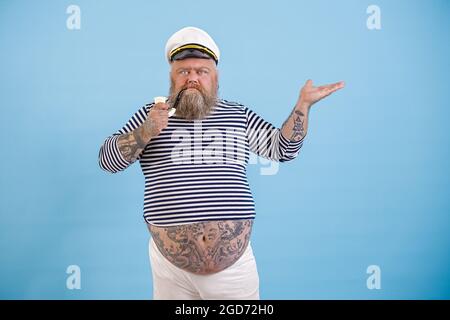 L'homme de la graisse en train de battre des tatouages en costume de marin pointe à côté sur fond bleu clair Banque D'Images