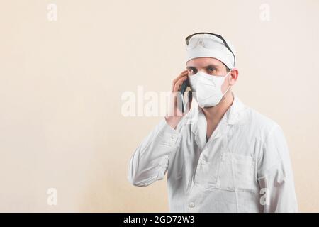 Jeune médecin en blouse de laboratoire blanche, lunettes de protection et masque respiratoire de protection tenant le téléphone cellulaire parler par mobile isolé. Banque D'Images