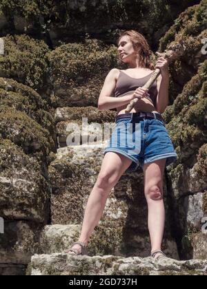 Une fille tenant un bois flotté sous forme de club se tient sur les rochers surcultivés avec de la mousse et illuminés par le soleil. Enregistrer l'image Banque D'Images