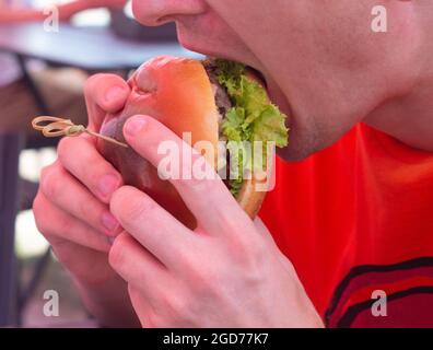 Un homme mange un gros hamburger juteux dans un magasin de hamburgers. Hamburger avec fromage, légumes, côtelettes gros plan. Banque D'Images