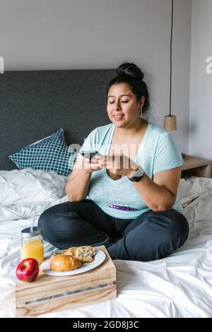 Jeune femme mexicaine de plus grande taille assise au lit à l'aide du téléphone tout en prenant le petit déjeuner en Amérique latine Banque D'Images