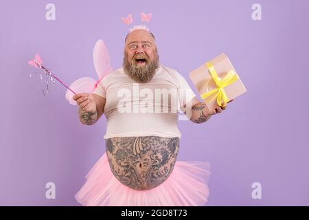 Joyeux homme obèse en costume de fée tient boîte cadeau et bâton magique sur fond violet Banque D'Images