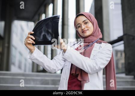 Gros plan sur le portrait d'une femme musulmane souriante médecin tenant une radiographie. Jolie charmante femme arabe médecin ou infirmière, portant un manteau médical, debout à l'extérieur du bâtiment moderne avec radiographie Banque D'Images