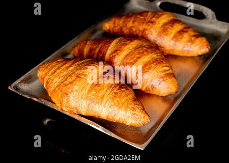 Trois croissants maison parfumés sur un plateau métallique, en gros plan, isolés sur du noir. Banque D'Images