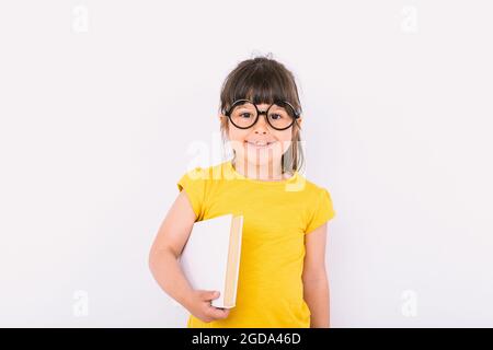 Petite fille souriante portant un t-shirt jaune et des lunettes noires rondes tenant un livre dans ses mains sur fond blanc Banque D'Images