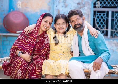 Portrait d'une famille indienne rurale heureuse en regardant l'appareil-photo tout en étant assis sur un lit traditionnel à la maison du village, adorable petite fille câlin sa graisse Banque D'Images