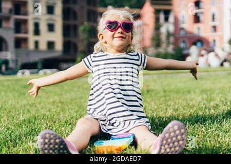 Une petite blonde drôle de lunettes de soleil et une robe rayée se trouve sur la pelouse. Banque D'Images