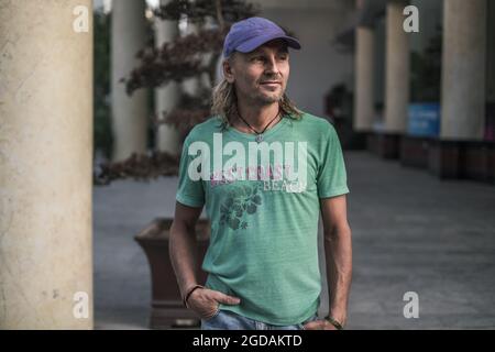 Homme mûr avec de longs cheveux gris portant un t-shirt vert et une casquette bleue debout sur la rue en ville et regardant de côté. Portrait. Photo de haute qualité Banque D'Images