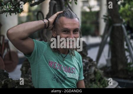 Homme mûr souriant avec de longs cheveux gris portant un t-shirt vert assis dans la rue en ville et tenant ses cheveux. Portrait. Photo de haute qualité Banque D'Images