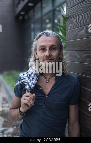 Homme mûr et beau en t-shirt et jeans debout à l'extérieur tenant sa chemise dans la main. Portrait en gros plan. Style grunge. Bâtiment moderne sombre Banque D'Images