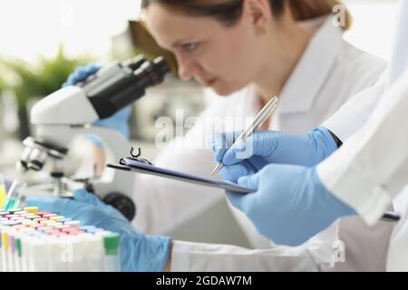 Femme chimiste regardant au microscope en laboratoire. Assistant écrivant les données sur papier Banque D'Images